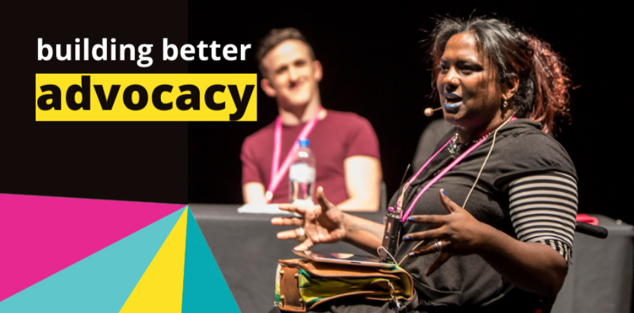 A woman is setting in a wheelchair on a stage. She is wearing a microphone headset and is speaking into it. Behind her are two people sitting at a table. The words BUILDING BETTER ADVOCACY are in the top left hand corner of the image. A graphic element comprising pink, yellow and blues shapes is in the bottom left corner.