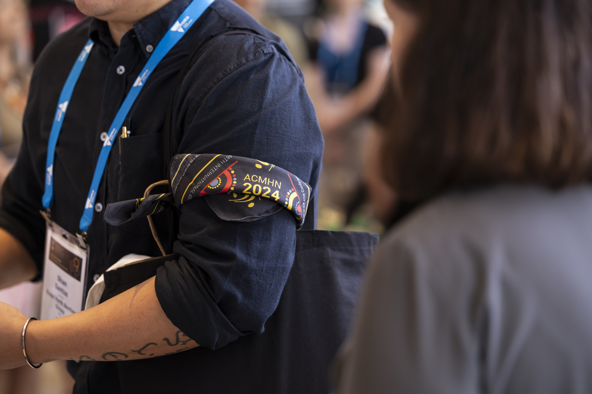 A person wearing a folded ACMHN 2024 bandana over their arm.